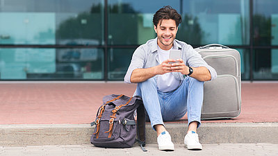 Reisender Mann sitzt mit Smartphone und Koffer am Gehsteig vor Flughafenhalle