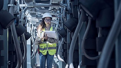 Foto: Technikerin mit Schutzhelm, Schutzbrille und Warnweste steht zwischen technischen Anlagen und Kabelsystemen und hält ein Klemmbrett zur Kontrolle von Infrastrukturkomponenten.