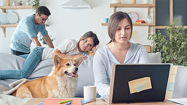 Frau arbeitet im Homeoffice am Laptop mit Tochter, Hund und Mann im Hintergrund