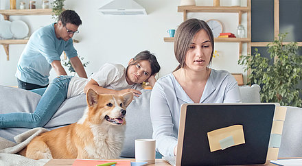 Frau arbeitet im Homeoffice am Laptop mit Tochter, Hund und Mann im Hintergrund